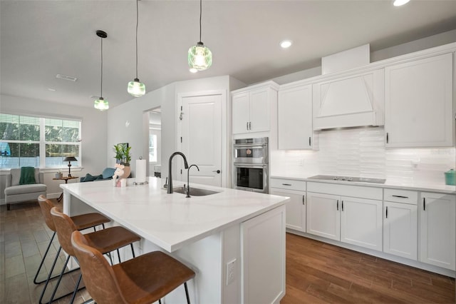 kitchen featuring stainless steel double oven, dark hardwood / wood-style flooring, an island with sink, pendant lighting, and white cabinets