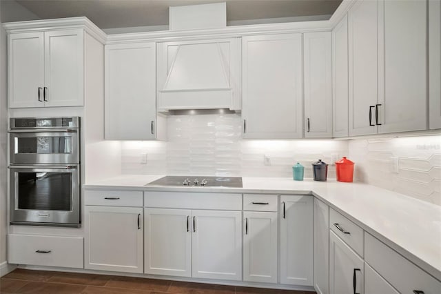 kitchen with backsplash, black cooktop, custom exhaust hood, stainless steel double oven, and white cabinets