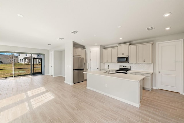 kitchen with appliances with stainless steel finishes, tasteful backsplash, sink, a center island with sink, and light hardwood / wood-style flooring