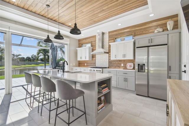 kitchen featuring a raised ceiling, an island with sink, wall chimney range hood, and appliances with stainless steel finishes