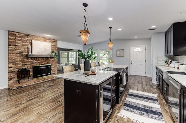 kitchen featuring a brick fireplace, electric range, hanging light fixtures, an island with sink, and backsplash