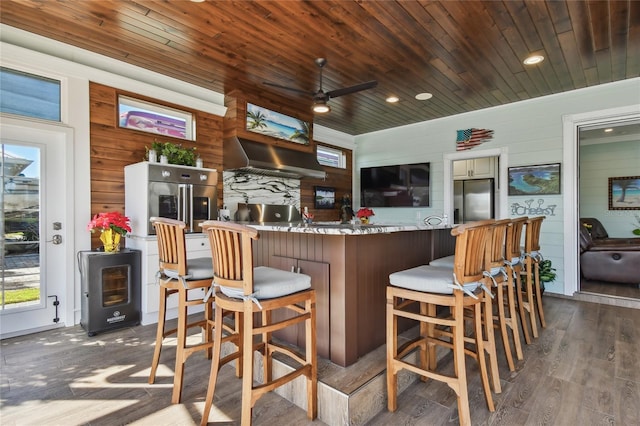 kitchen featuring dark wood-type flooring, wall chimney range hood, wooden walls, stainless steel fridge with ice dispenser, and wood ceiling