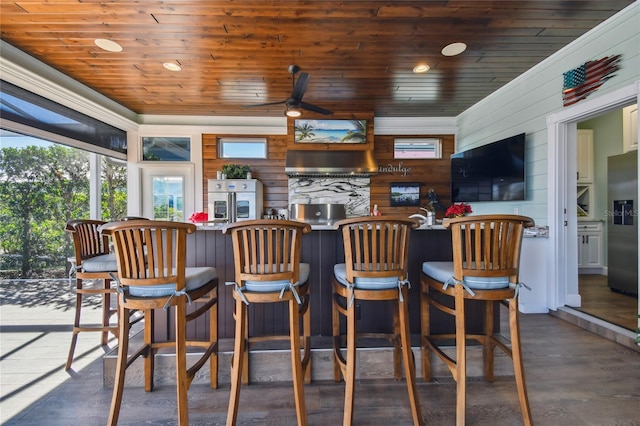 bar with stainless steel fridge with ice dispenser, ceiling fan, wooden ceiling, and wall chimney range hood