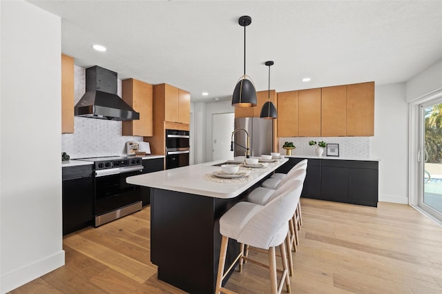 kitchen with wall chimney exhaust hood, stainless steel appliances, pendant lighting, light hardwood / wood-style flooring, and an island with sink