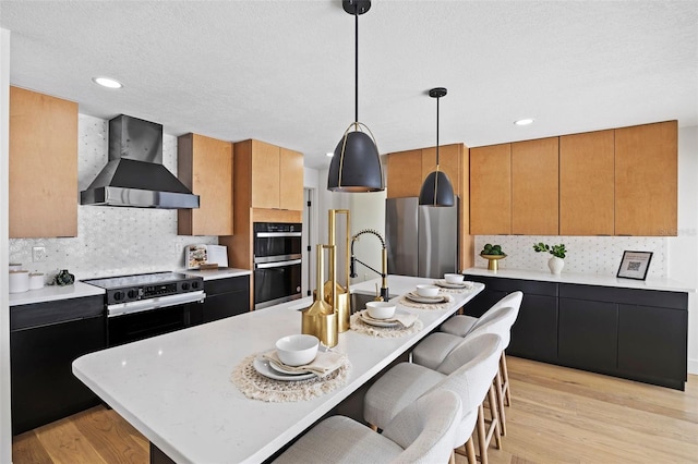 kitchen featuring pendant lighting, wall chimney range hood, light wood-type flooring, an island with sink, and appliances with stainless steel finishes