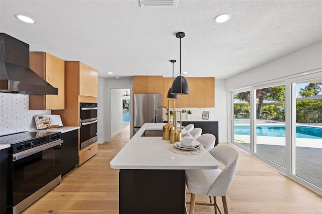 kitchen featuring stainless steel appliances, a kitchen island with sink, pendant lighting, light hardwood / wood-style flooring, and range hood