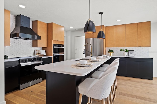 kitchen with decorative light fixtures, a center island with sink, stainless steel appliances, and wall chimney range hood