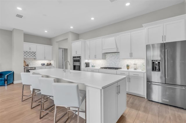 kitchen with white cabinetry, stainless steel appliances, backsplash, a center island with sink, and custom exhaust hood