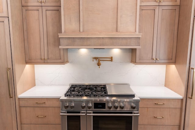 kitchen featuring custom exhaust hood, light brown cabinets, range with two ovens, decorative backsplash, and light stone counters