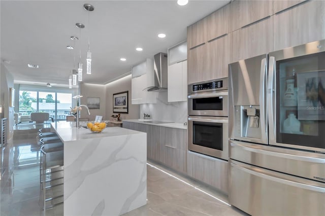 kitchen with hanging light fixtures, stainless steel appliances, wall chimney range hood, an island with sink, and a breakfast bar area