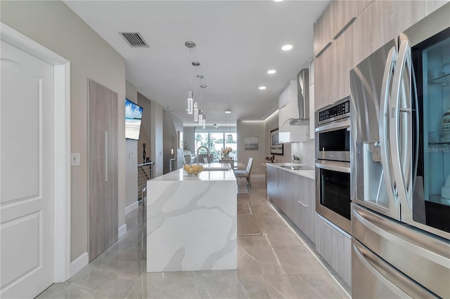 kitchen featuring light stone countertops, appliances with stainless steel finishes, wall chimney range hood, a center island with sink, and hanging light fixtures