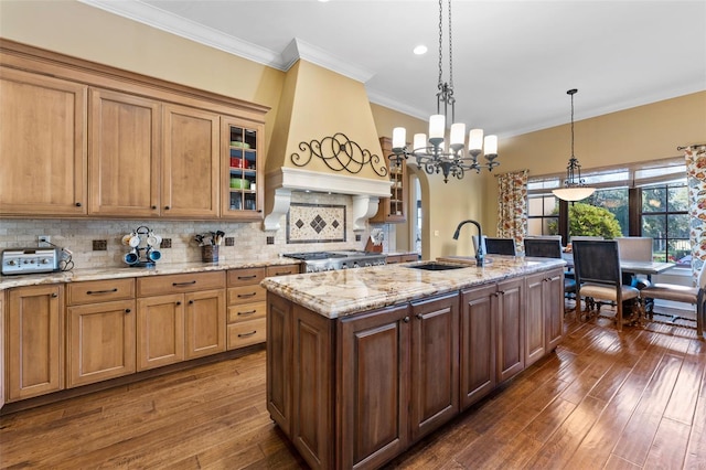 kitchen with dark hardwood / wood-style flooring, premium range hood, decorative light fixtures, a kitchen island with sink, and ornamental molding