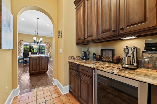 bar featuring light wood-type flooring, light stone counters, ornamental molding, beverage cooler, and an inviting chandelier