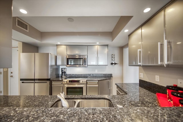 kitchen with appliances with stainless steel finishes, gray cabinetry, a tray ceiling, sink, and dark stone countertops