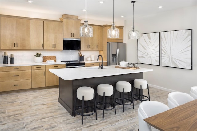 kitchen with appliances with stainless steel finishes, pendant lighting, a kitchen island with sink, and light brown cabinetry