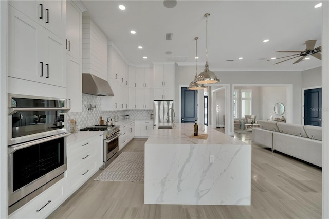 kitchen with appliances with stainless steel finishes, ventilation hood, a kitchen island with sink, white cabinets, and hanging light fixtures