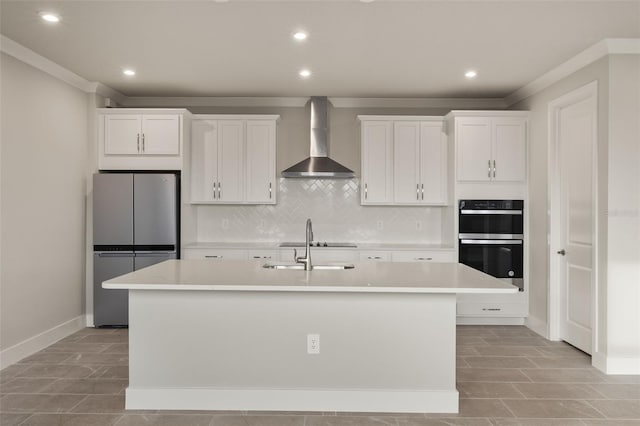 kitchen featuring wall chimney exhaust hood, stainless steel refrigerator, double oven, an island with sink, and white cabinets