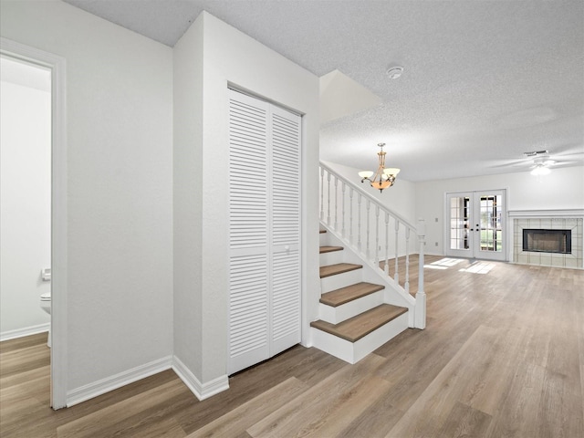 stairway with french doors, wood-type flooring, a textured ceiling, a tiled fireplace, and ceiling fan with notable chandelier