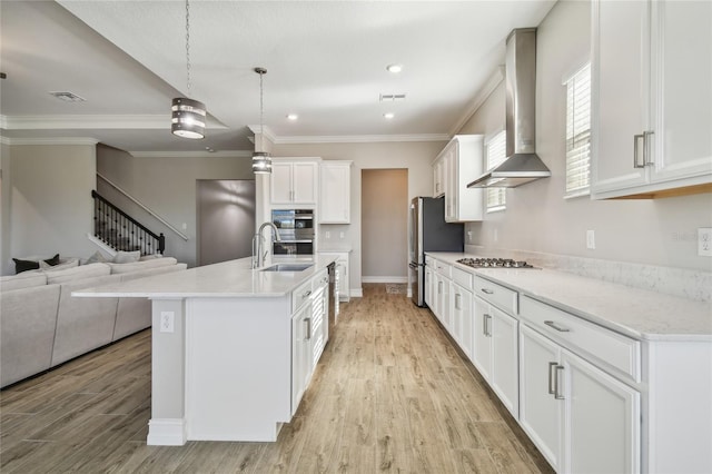 kitchen featuring light wood-type flooring, wall chimney exhaust hood, decorative light fixtures, white cabinetry, and an island with sink