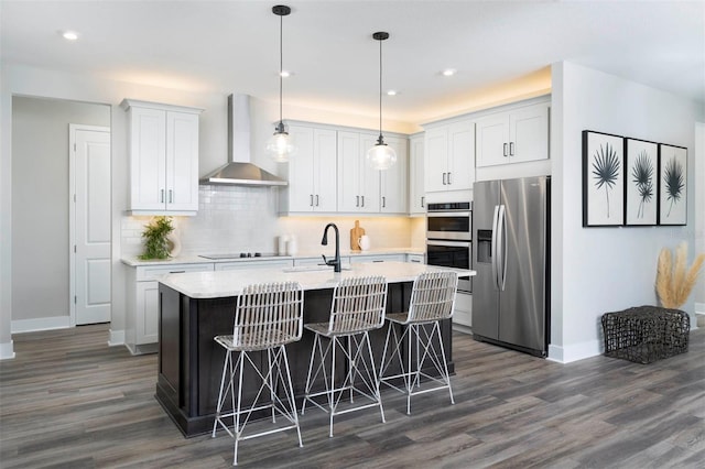 kitchen with wall chimney exhaust hood, stainless steel appliances, a kitchen island with sink, dark wood-type flooring, and white cabinetry