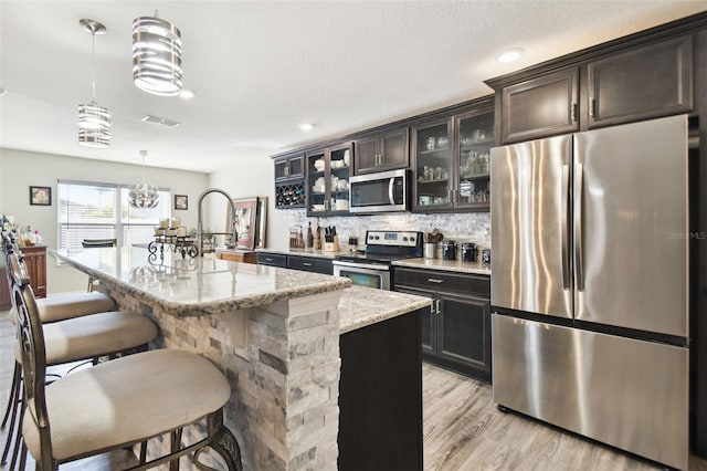 kitchen featuring light stone countertops, dark brown cabinets, stainless steel appliances, a kitchen island with sink, and hanging light fixtures