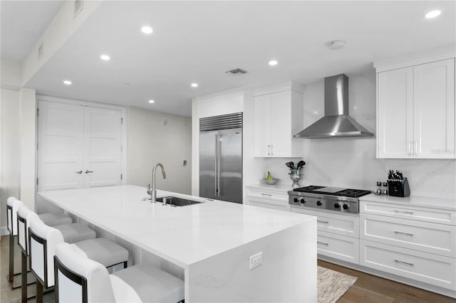 kitchen featuring wall chimney range hood, sink, an island with sink, appliances with stainless steel finishes, and white cabinetry