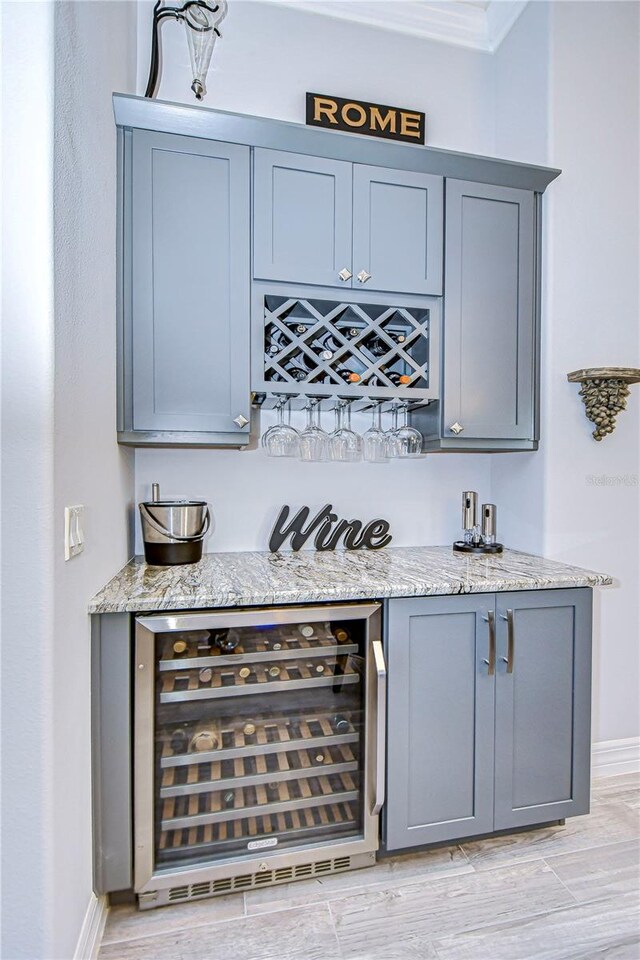 bar with light stone counters, ornamental molding, wine cooler, and light hardwood / wood-style flooring