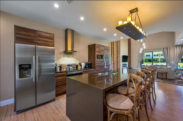 kitchen with sink, wall chimney exhaust hood, built in appliances, light hardwood / wood-style floors, and a kitchen island with sink