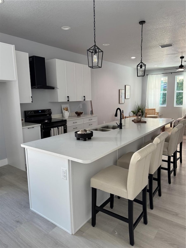 kitchen featuring black range with electric cooktop, a kitchen island with sink, a sink, white cabinets, and wall chimney range hood