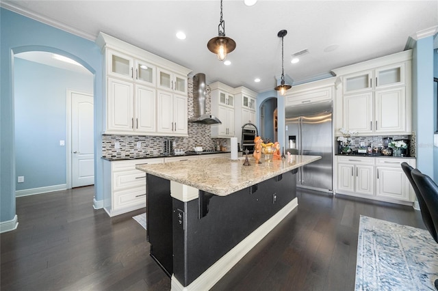 kitchen featuring white cabinetry, hanging light fixtures, stainless steel built in refrigerator, an island with sink, and wall chimney exhaust hood