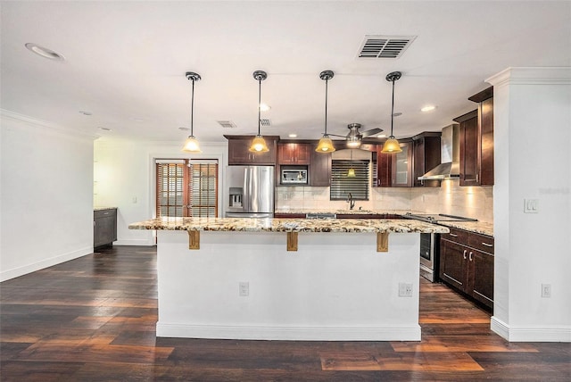 kitchen featuring a center island, wall chimney range hood, hanging light fixtures, light stone countertops, and stainless steel appliances