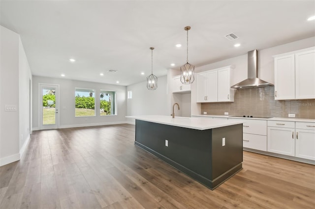 kitchen featuring decorative light fixtures, tasteful backsplash, white cabinetry, an island with sink, and wall chimney range hood