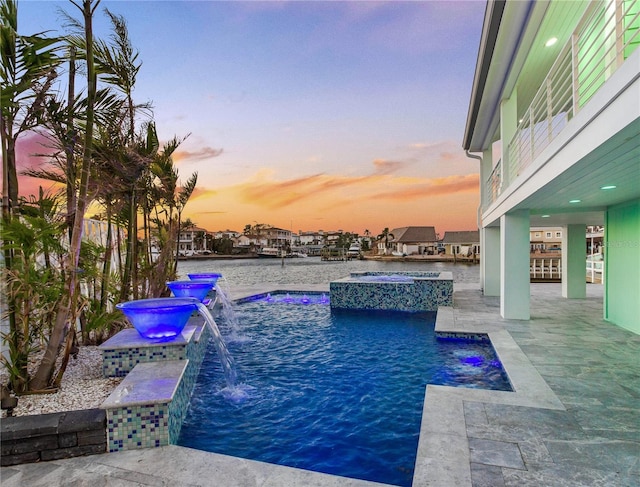 pool at dusk with a water view, an in ground hot tub, pool water feature, and a patio