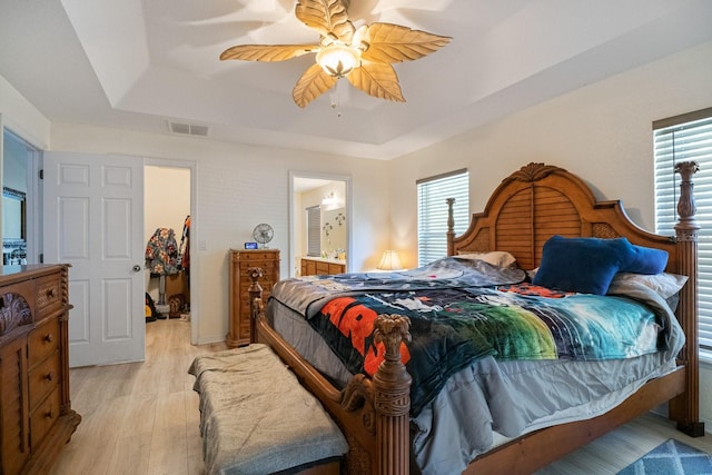 bedroom featuring ceiling fan, light wood-type flooring, connected bathroom, and a tray ceiling