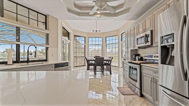 kitchen with light stone countertops, stainless steel appliances, a tray ceiling, sink, and hanging light fixtures