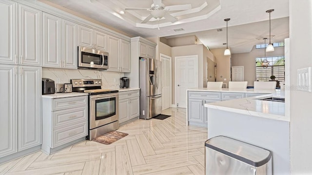 kitchen featuring light stone counters, stainless steel appliances, a raised ceiling, ceiling fan, and decorative light fixtures