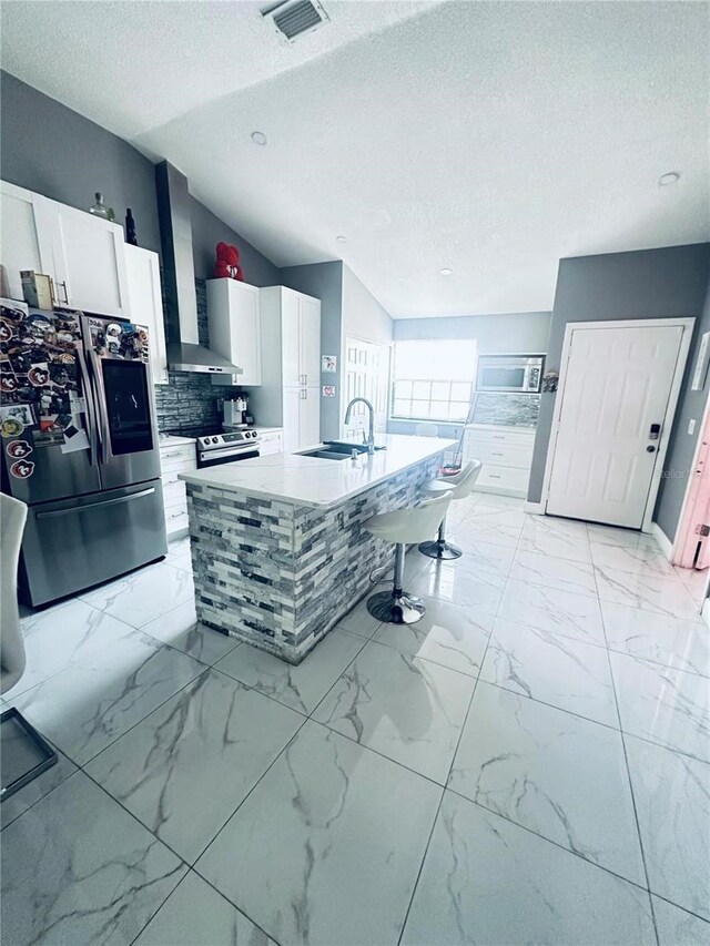 kitchen with wall chimney exhaust hood, stainless steel appliances, an island with sink, a textured ceiling, and white cabinets