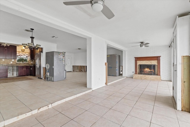 unfurnished living room with light tile patterned floors, ceiling fan with notable chandelier, a brick fireplace, and brick wall