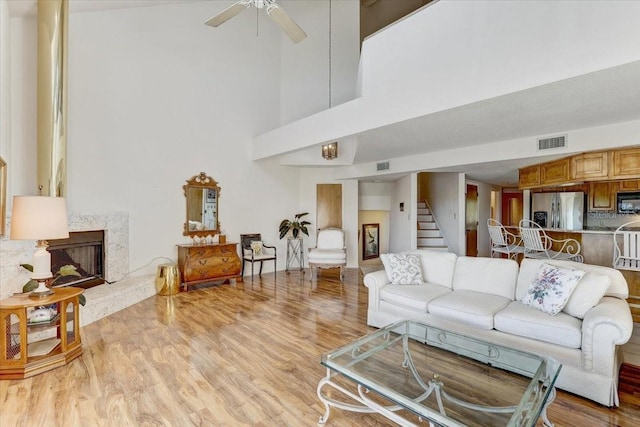 living room with a towering ceiling, light wood-type flooring, ceiling fan, and a premium fireplace