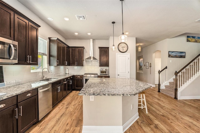 kitchen with sink, stainless steel appliances, wall chimney range hood, a kitchen island, and light wood-type flooring