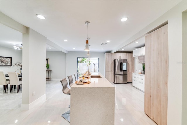 kitchen with a kitchen island with sink, sink, pendant lighting, white cabinetry, and stainless steel refrigerator