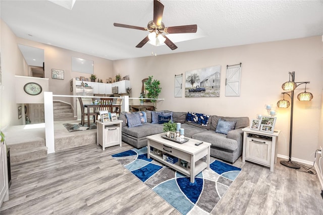 living area featuring light wood finished floors, ceiling fan, stairs, baseboards, and a textured ceiling