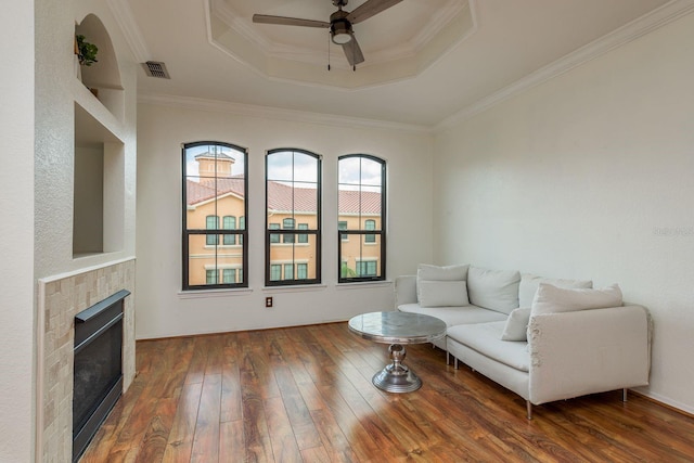 living room with ornamental molding, a tray ceiling, ceiling fan, dark wood-type flooring, and a tiled fireplace