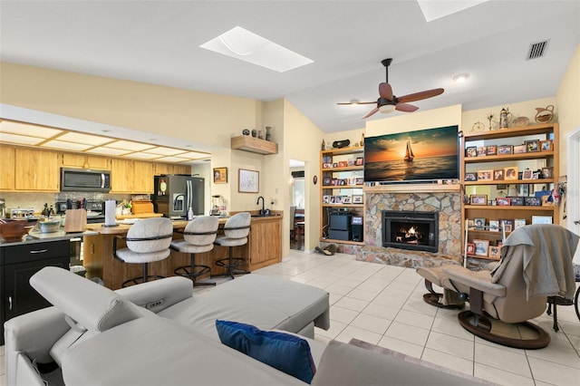 tiled living room with vaulted ceiling with skylight, ceiling fan, and a stone fireplace