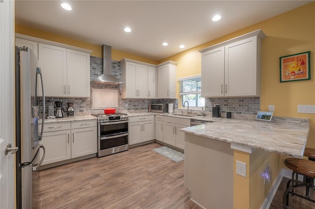 kitchen featuring kitchen peninsula, appliances with stainless steel finishes, a breakfast bar, wall chimney range hood, and white cabinetry