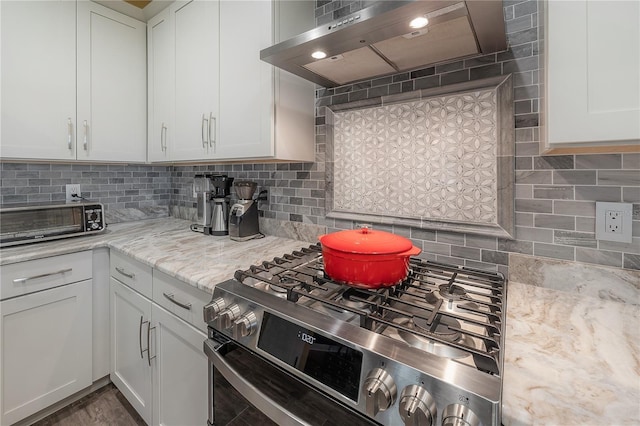 kitchen with light stone countertops, wall chimney exhaust hood, stainless steel gas range oven, backsplash, and white cabinets