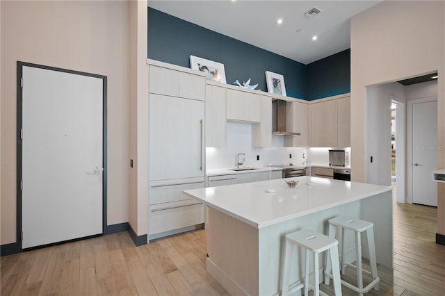 kitchen with sink, wall chimney exhaust hood, light wood-type flooring, a kitchen island, and white cabinetry