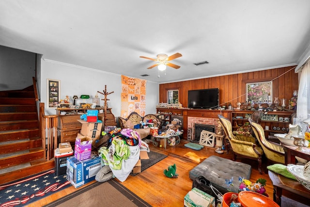 living room featuring wooden walls, a brick fireplace, ceiling fan, ornamental molding, and wood-type flooring