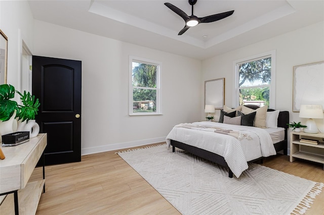 bedroom featuring a tray ceiling, multiple windows, ceiling fan, and light wood-type flooring