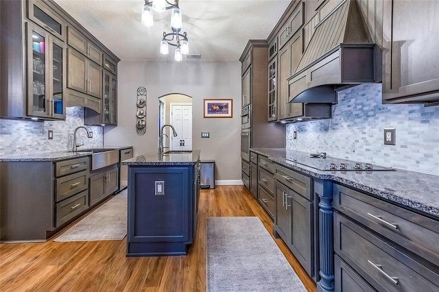 kitchen with black electric stovetop, light hardwood / wood-style floors, dark stone counters, and an island with sink